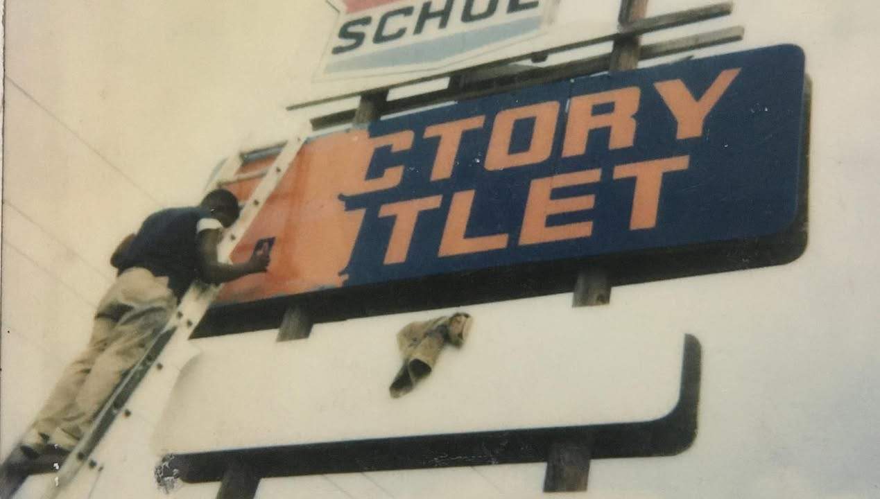 A Butler Signs craftsman repainting a factory outlet storefront sign from a ladder
